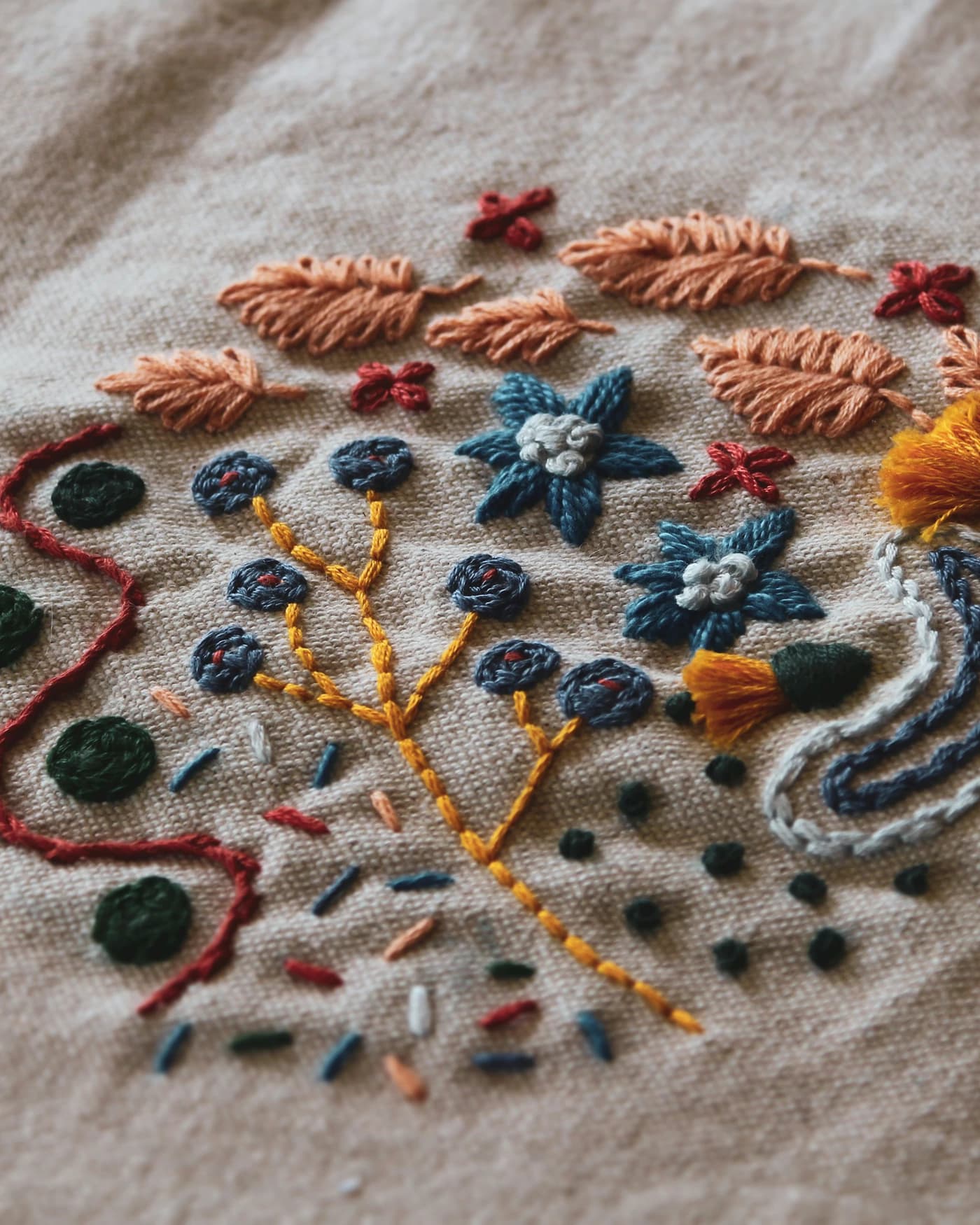 Close-up of hand embroidery on natural linen — raised floral stitchwork in rust, indigo and gold.