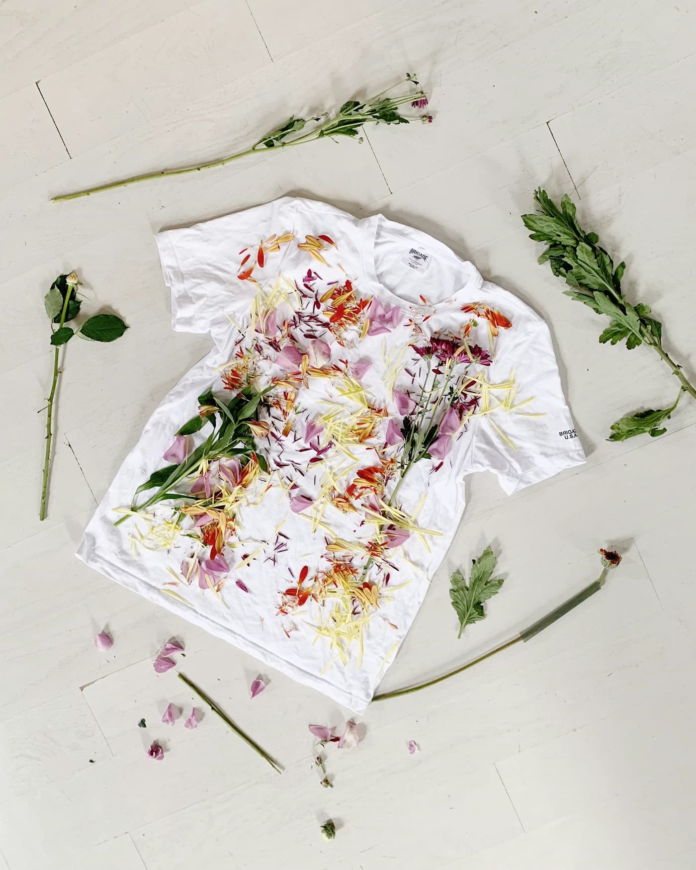 A white tee laid flat, its front covered in a loose painted floral print, cut stems arranged around it.