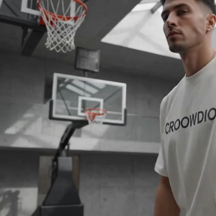 A man in a bone-white CROOWDIO tee, seen from below against a concrete stand and hoop.