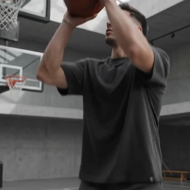 A player raising a basketball to shoot, the sleeves of his charcoal tee falling loose.