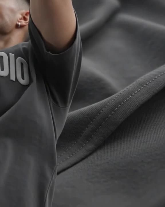 A raised arm pulling the shoulder of a heavyweight charcoal tee taut, the fold and shoulder seam catching the light.