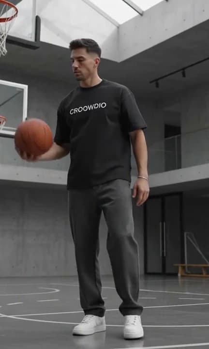 A man in an oversized black CROOWDIO tee and charcoal trousers standing on an empty indoor court, a basketball at his hip.